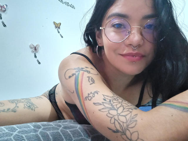 EmmaRuiz with tattoos, wearing round glasses, black bra, and hoop earrings, has a nose ring and lip piercing, lying on a grey-patterned blanket, smiling at the camera.