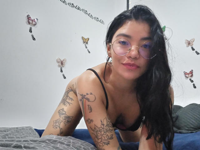 EmmaRuiz, with tattoos on her arms, leaning forward in black lingerie, wearing glasses and a nose ring, has pink lips and black hair, on a blue bed.