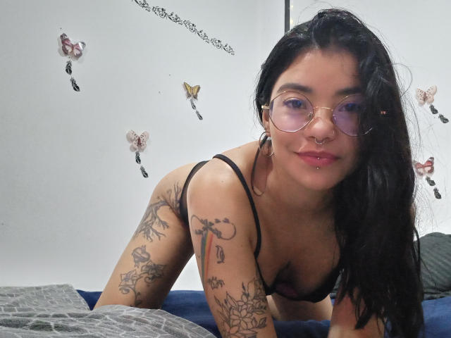 EmmaRuiz with long black hair, glasses, nose ring, and lip piercing, leaning on bed in black bra, showcasing tattoos on arms and chest, smiling at camera.
