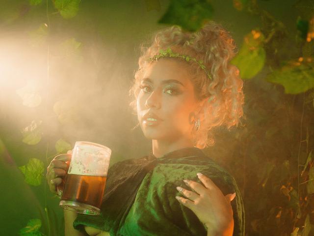 LilithWalker with curly blonde hair, green leaf headband, and green dress, holding a beer, sunlight on her face, green leaves in the background, looking slightly to the side.