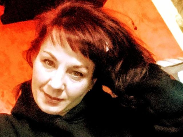 DianaAhTender ha i capelli rossi, la pelle chiara e sta sorridendo alla fotocamera. Indossa un maglione nero a collo alto e la sua testa è leggermente inclinata verso destra..