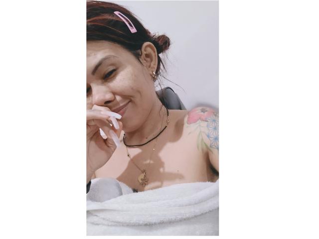 NathashaRomexo con cabello castaño oscuro recogido en un moño, pinza rosa en el cabello, sonriendo, mano en la barbilla, toalla blanca, tatuaje en el hombro, collar, uñas blancas, senos desnudos, relajada..