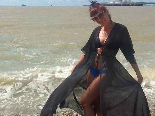 Diana, cheveux bruns attachés en chignon, portant une robe noire transparente, un bikini bleu, des lunettes de soleil et un bandeau noir, debout dans de l'eau peu profonde, montrant ses seins et ses fesses..