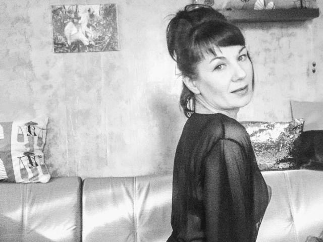 Diana, les cheveux noirs attachés en chignon, portant un haut noir transparent, regardant en arrière avec un léger sourire, montrant ses seins, assise sur un canapé en cuir brillant..