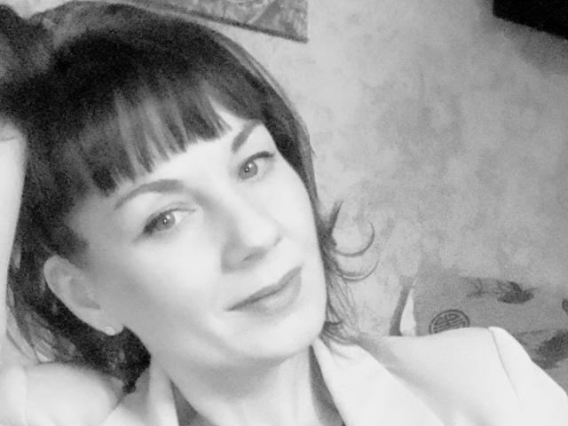 Black-and-white selfie of DianaAhTender with dark bangs, light skin, and a subtle smile, showing her face and upper chest, wearing a light-colored shirt, hand in her hair.