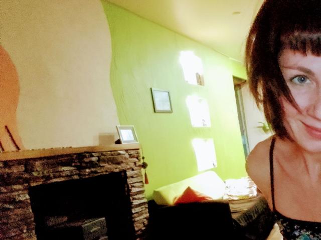 DianaAhTender with short brown hair and blue eyes, smiling, wearing a black spaghetti strap top, showing her shoulders and upper chest, standing in a room with a stone fireplace.