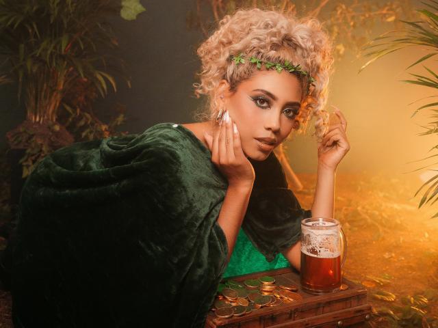 LilithWalker with curly blonde hair, green leaf headband, green velvet cloak, looking at camera, holding her chin, next to a glass of beer with foam, on a gold-covered surface.