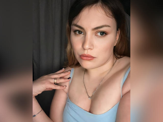 AnnabellkaSecret with light brown eyes, full lips, and medium tits in a blue tank top, touching her chest, wearing a necklace, and a ring on her finger.