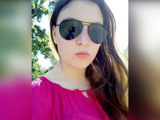 AnnabellkaSecret with long brown hair, wearing dark round sunglasses, and a pink top, has a serious expression, and light skin, standing outdoors in bright sunlight.