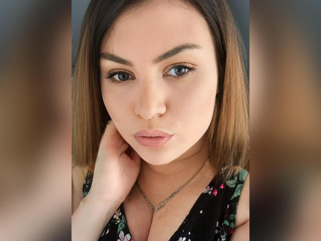 AnnabellkaSecret has light brown skin, straight brown hair, and is looking at the camera with a slight pout, wearing a floral dress, her hand near her chin.