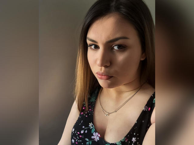 AnnabellkaSecret with light brown skin, medium-length dark brown hair with blonde highlights, wearing a floral black dress, small tits, subtle makeup, and a gold necklace, looking serious.