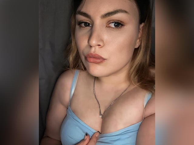 AnnabellkaSecret with light brown hair, blue eyes, and full lips, wearing a light blue tank top, showing her tits, slightly parted, with a hand on her chest.