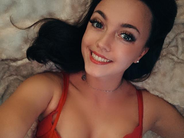 Vanilia69 with dark hair, red lingerie, smiling, showing teeth, blue eyes, makeup, necklace, lying on beige blanket, upper body, cute, sexy.