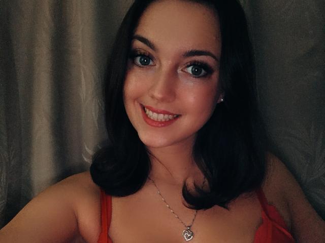 Vanilia69 has dark brown hair, blue eyes, and a cute smile with red lipstick. She's wearing a red top and a silver necklace with a heart-shaped pendant.