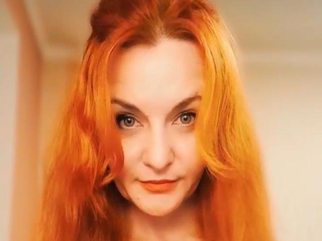 JasminsLove with orange-red hair, looking up with a slight smile, has blue eyes and light skin, wearing orange lipstick, in a soft, warm-lit selfie.