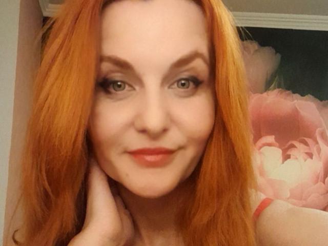 JasminsLove mit roten Haaren, grünen Augen und einem süßen Lächeln, die sich ihre Wange hält, trägt einen roten Träger und zeigt ihr Gesicht in einem Selfie..