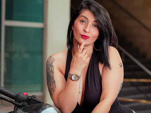 KarenBroc, with black hair, red lipstick, and a black halter top, shows a finger to her lips, wearing a watch, and has tattoos on her arm and shoulder.