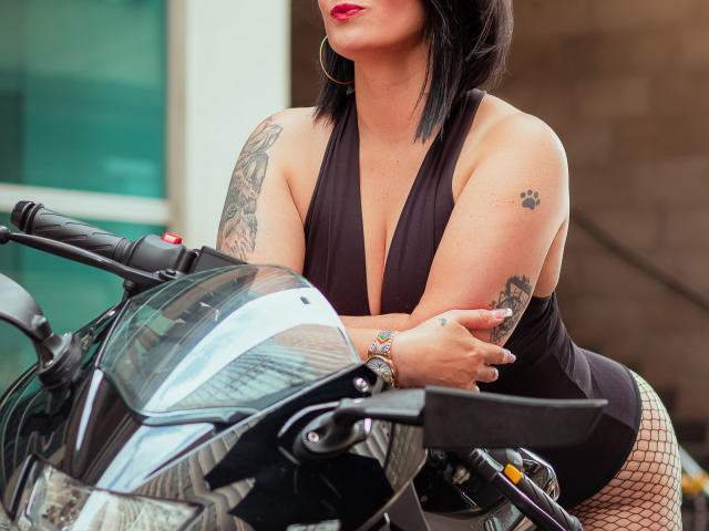 KarenBroc with black bob, red lipstick, sleeve tattoos, and fishnet stockings, leaning on a black motorcycle, wearing a deep V black dress, showing her tits and ass.