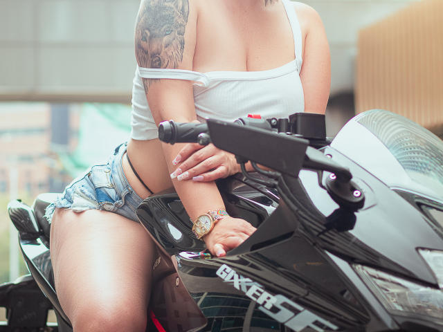 KarenBroc with a tattooed arm, white off-shoulder top, and denim shorts, leaning on a black "BAXERER" motorcycle, showing her tits and wearing a colorful wristwatch.