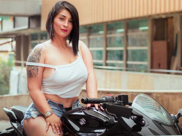 KarenBroc with black bob, white tank top, and denim shorts, sitting on a black motorcycle, showing her big tits and tattooed arm, wearing a watch, smiling.