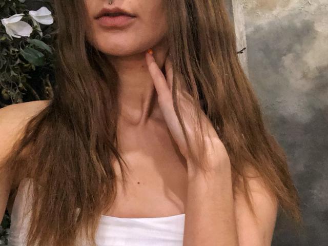 Karish's face, showing brown hair, a nose piercing, and her hand touching her neck with orange-painted nails, wearing a white strap top, exposing her small tits.