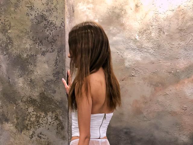 Karish with long brown hair, back to the camera, wearing a white strapless top with a zipper, standing against a textured wall, her hands touching the wall.