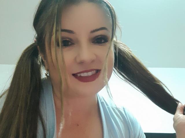 NatalyHami mit voluminösem, braunem Haar zu Zöpfen, trägt ein hellblaues Oberteil, rote Lippenstift, lächelt und hält eine Haarsträhne, im Hintergrund ist eine weiße Decke zu sehen..