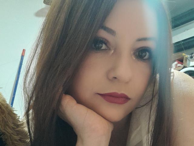 NatalyHami with brown hair, red lipstick, and dark eye makeup, resting her chin on her hand, looking at the camera with a slightly bored expression.