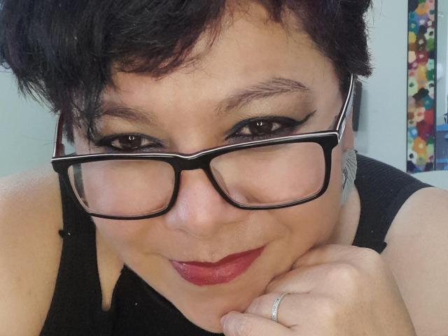Angellovelly has short black hair, wearing thick black glasses, and red lipstick. She's smiling with her chin on her hand, showing a ring on her finger.