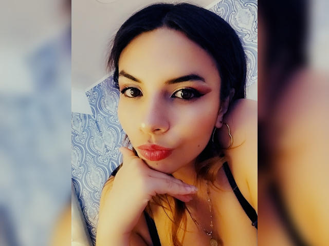 VioletaStone with dark hair, red lipstick, and hoop earrings, pouting with hand on chin, wearing black bra, reflected in mirror, showing makeup and earrings.