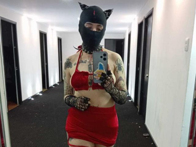 AmySpider in a red bikini top and skirt, black cat mask, tattoos, fishnet gloves, holding phone in hallway. Boobs visible, red ribbon tied to skirt, sexy look.