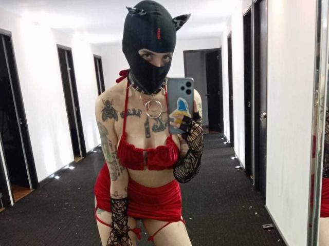 AmySpider in red lingerie with black fishnet arms, black cat mask, tattoos, and holding a phone, showing her tits and ass in a mirror selfie.