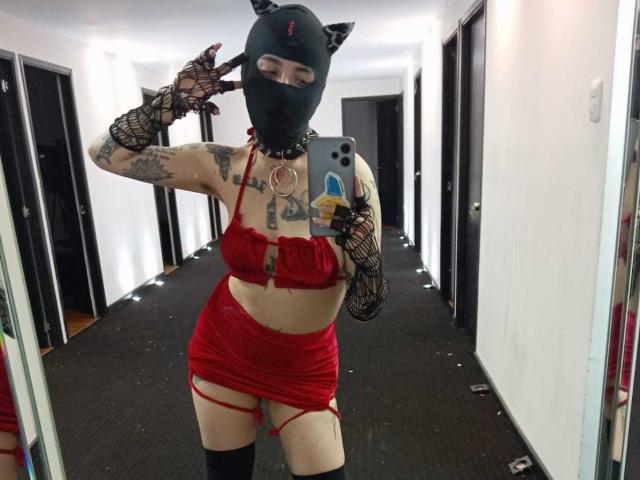 AmySpider indossa un set di lingerie rossa con guanti a rete nera, una maschera da gatto e tatuaggi, mostrando il seno e i glutei in un selfie allo specchio, indossando bretelle alte..