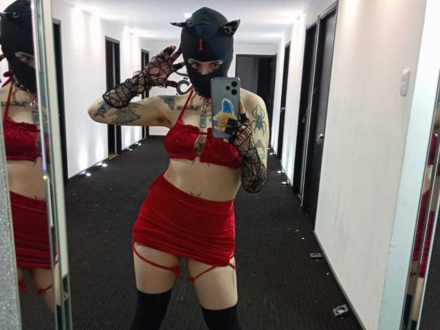 AmySpider, avec des tatouages, portant un masque noir, des oreilles de chat, un soutien-gorge en dentelle rouge, une jupe en dentelle rouge, des gants en résille noire, des bas noirs, prenant un selfie devant un miroir dans un couloir..