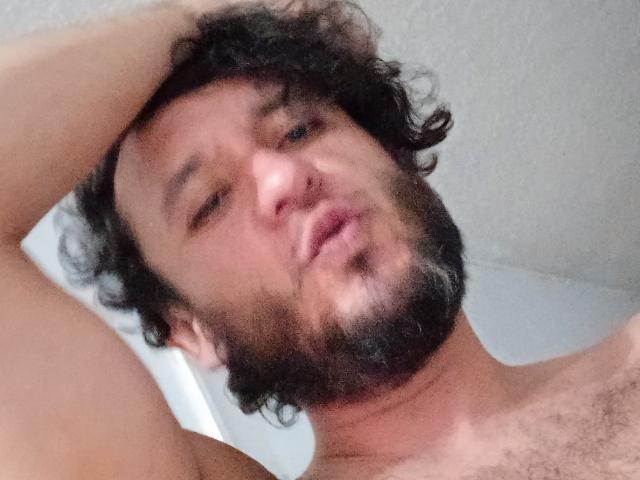 DavydWolfy69, con capelli neri ricci e disordinati, barba e leggera peluria, che fa l'occhiolino, alza il braccio destro, senza camicia, mostrando i peli del petto, in un selfie scattato leggermente di lato e da un'angolazione bassa..