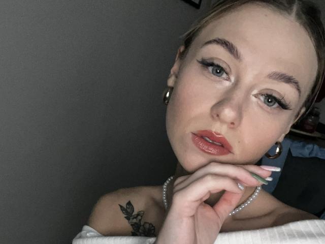 CindyxGlam has light skin, blue eyes, and glossy pink lips. She's wearing hoop earrings, a pearl necklace, and has a tattoo on her right shoulder. Her hand is near her face.