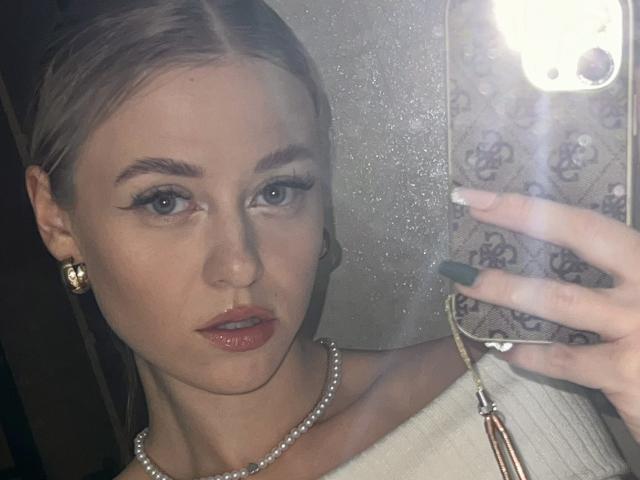 CindyxGlam with glossy pink lips, green nails, pearl necklace, gold earrings, and a white off-shoulder top, holding a phone with a floral patterned case in a mirror selfie.