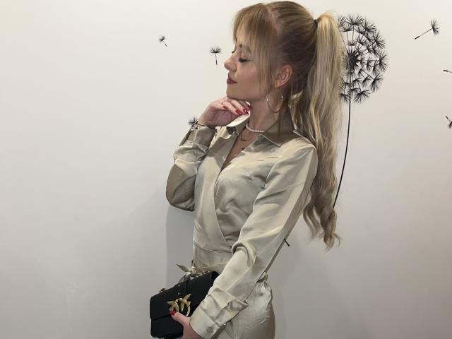 CindyxGlam mit blondem Pferdeschwanz, trägt ein beiges Hemd, schwarze Hose, eine zarte Halskette und steht vor einer weißen Wand mit einem großen schwarzen Löwenzahn. Die Brüste sind unter dem Hemd sichtbar. Die Hände sind in der Nähe des Gesichts..