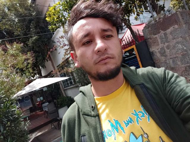 DavydWolfy69, avec un visage sérieux, des cheveux bruns courts, une veste verte, une chemise jaune et une légère barbe, se tient debout à l'extérieur en plein jour. Ses fesses ne sont pas visibles..