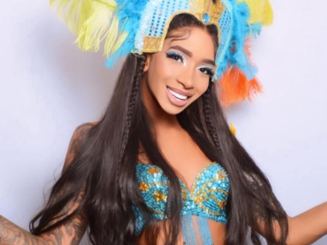 LunaBigCoock with long braids, wearing a colorful headdress and blue sequined bra, showing off her tits and smiling brightly. She has tattoos on her right arm.