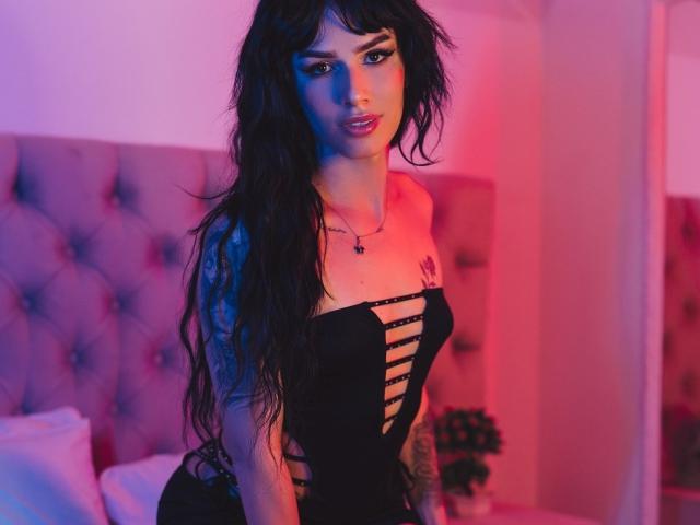 Maddox Carson, con cabello negro largo, vestida con un vestido negro sin tirantes con detalles recortados, iluminación rosa y azul, tatuajes en el brazo derecho y en el pecho, sentada en una cama..