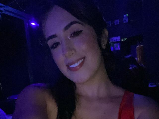 LaurenceRose with dark hair, smiling, wearing red top, black eyeliner, and nose piercing, blue-lit face, showing off a cute, sexy look. Her tits are partially visible.