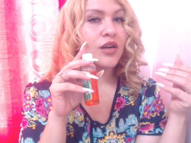 BigDirtyBoobs69 with blonde curls, floral shirt, and white nails, holding orange bottle, looking at camera with slightly open mouth. Bright pink curtain in background.