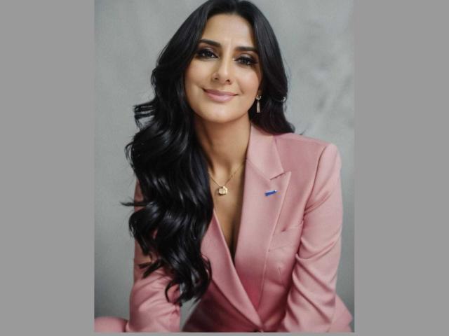 JuliaRobinson with long black wavy hair, pink suit, and blue pin, smiling at the camera, gold necklace, and subtle makeup, sitting against a gray background.