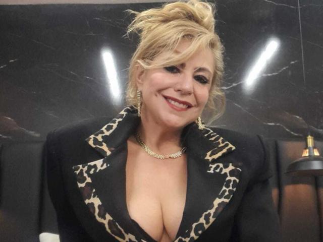 LylaGreen with blonde hair in a messy updo, smiling, wearing a black leopard-print blazer with a deep cleavage, gold necklace, and showing her large, round boobs.