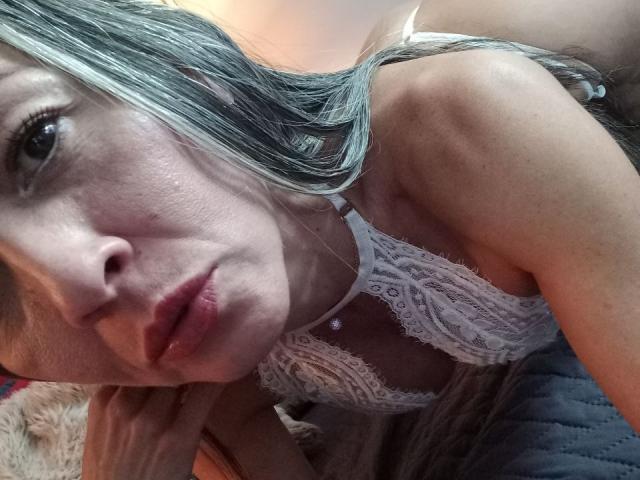 NicoletteShea's face with blue-gray hair, wearing a white lace bra, has puffy pink lips, slightly parted, with a hand on her cheek, showing her tits.