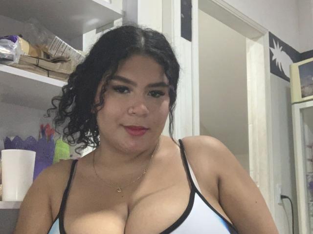 JulietaStark with curly black hair, red lipstick, and a gold necklace, wearing a white and black tank top, showing large tits. She's smiling slightly in a bedroom.