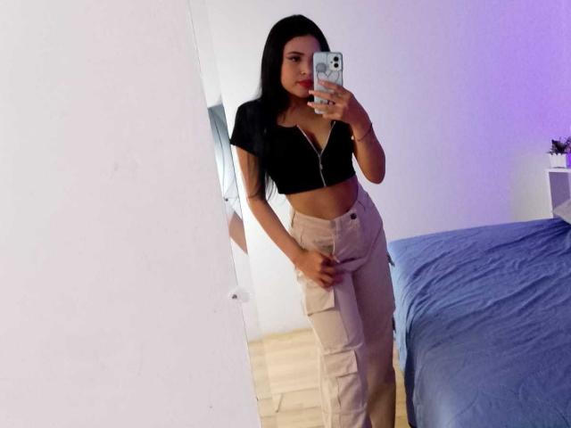 OliviGrace with long black hair, red lipstick, black crop top, and beige cargo pants, taking a mirror selfie, showing her tits and ass, standing in a room with a blue bed.