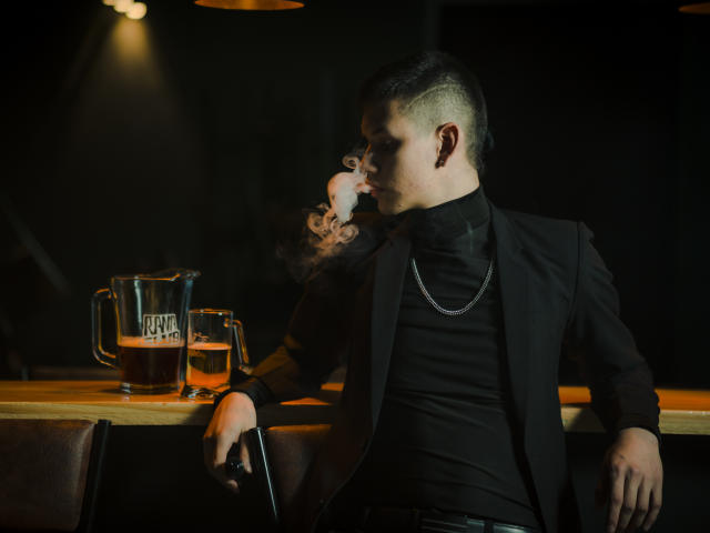 JeanKarloLeone with short, undercut hair, black suit, and chain, smoking, holding a black pen, leaning on a bar, with two beers in front of him.