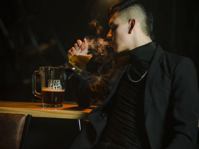 JeanKarloLeone with short undercut, black shirt, and jacket, smoking from a glass pipe, holding a beer mug, dark background, steam from pipe, looking to the side.